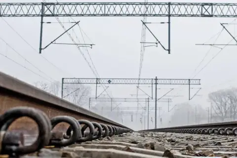 Torowisko kolejowe z bliska, z widocznymi śrubami mocującymi szyny. W tle sieć trakcyjna i mglisty krajobraz.