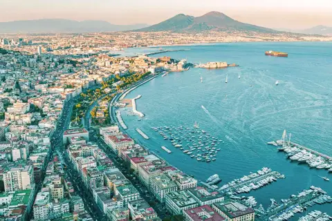 Widok na Neapol i Wezuwiusza. W oddali widać góry. W zatoce cumują łodzie. Można stąd ruszyć pociągiem do Sorrento.