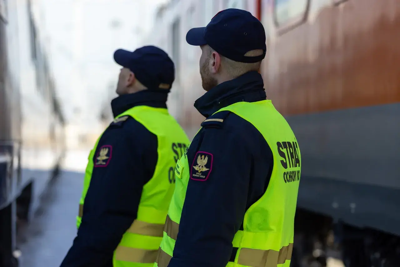 Dwóch funkcjonariuszy straży ochrony kolei w kamizelkach odblaskowych stoi przy pociągu, czuwając nad bezpieczeństwem.