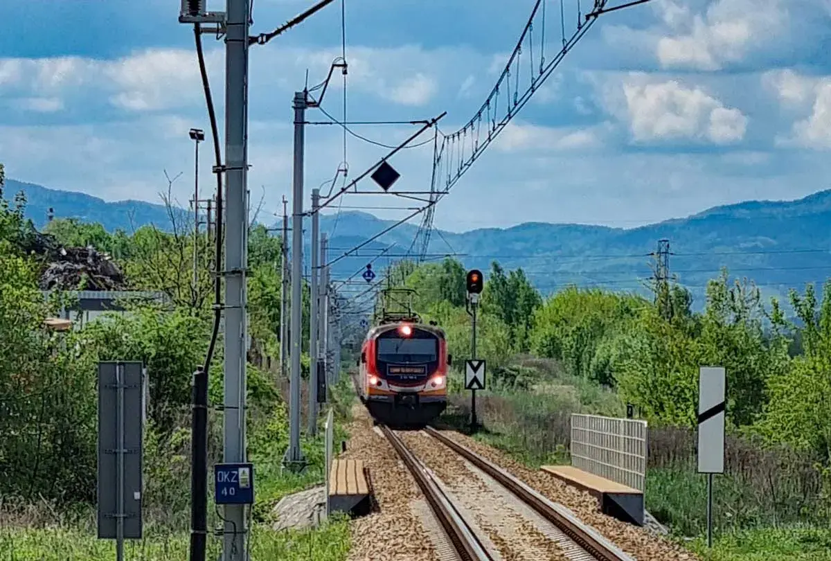 Pociąg PKP Łódź Kaliska - Krynica-Zdrój zbliża się do stacji, mijając zielone wzgórza i słupy trakcyjne.
