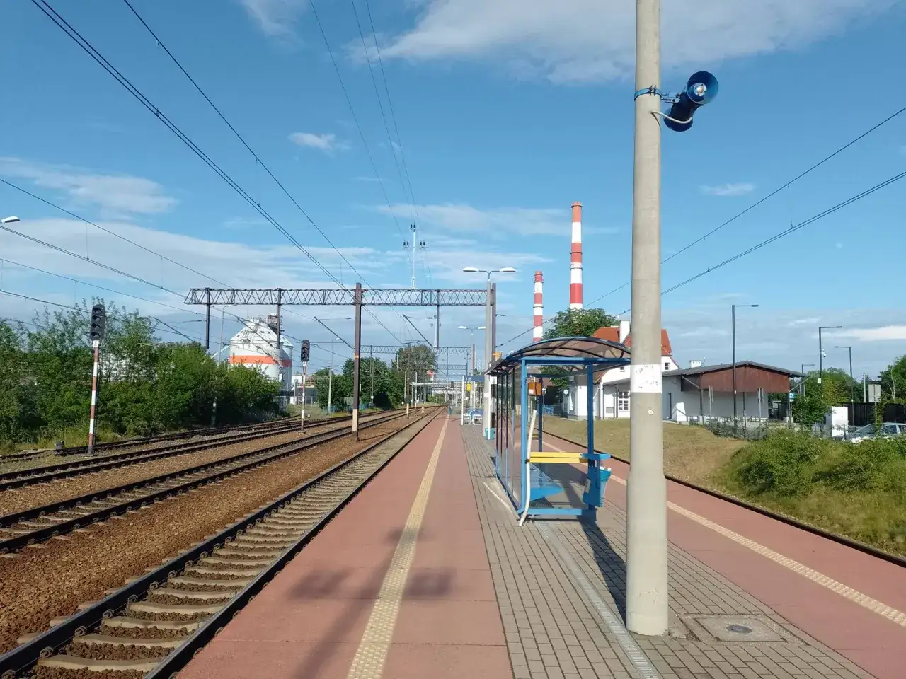 PKP Siechnice Wrocław: peron z wiatą, tory kolejowe i kominy w tle.